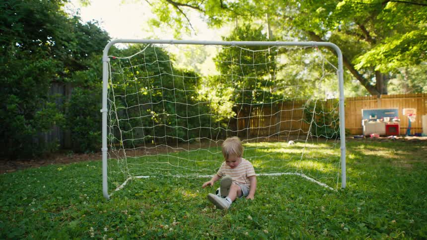 Playful toddler sits in a soccer net in a green backyard and holds a soccer ball up to his head