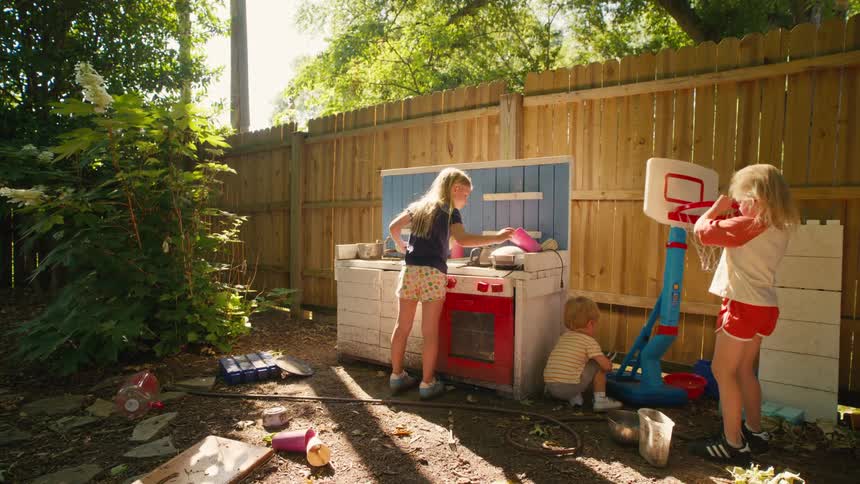 Three young children enjoy a sunny afternoon playing with their toy kitchen and basketball hoop in a messy backyard