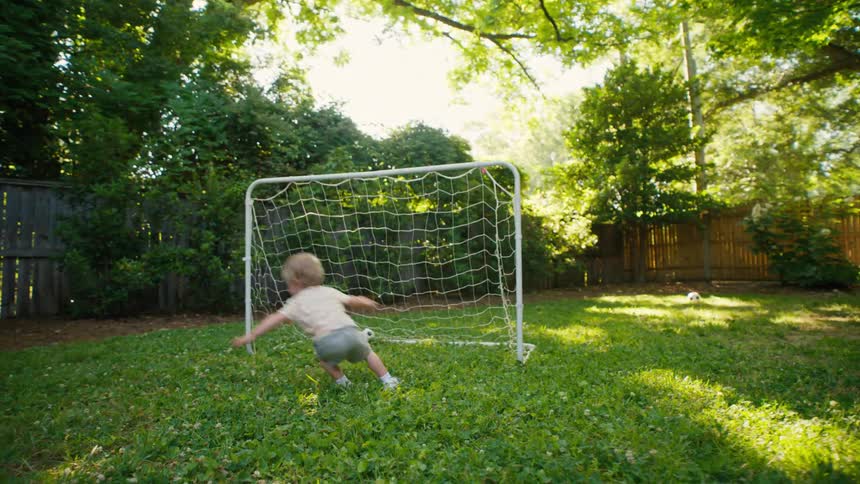 In a sunny backyard, a little boy joyfully plays soccer, running and kicking the ball before making a dramatic dive
