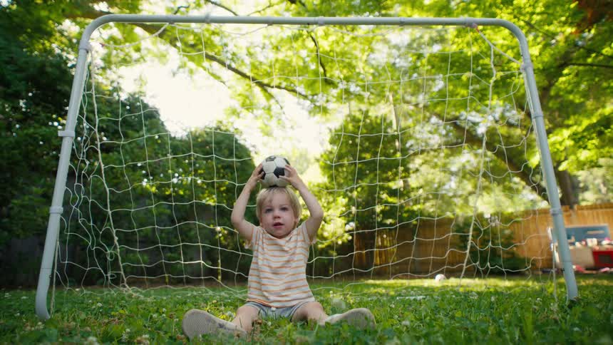 A happy toddler sits in a backyard soccer goal, playfully balancing a small soccer ball on his head and making funny faces