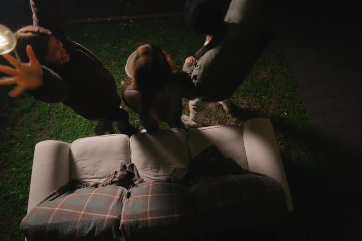 High-angle slow-motion shot of three energetic friends jumping up and dancing wildly on a couch in their backyard at night