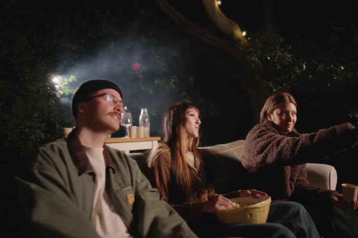 A group of young friends gather for a cozy backyard movie night, watching a film on a projector while sitting on a couch under string lights