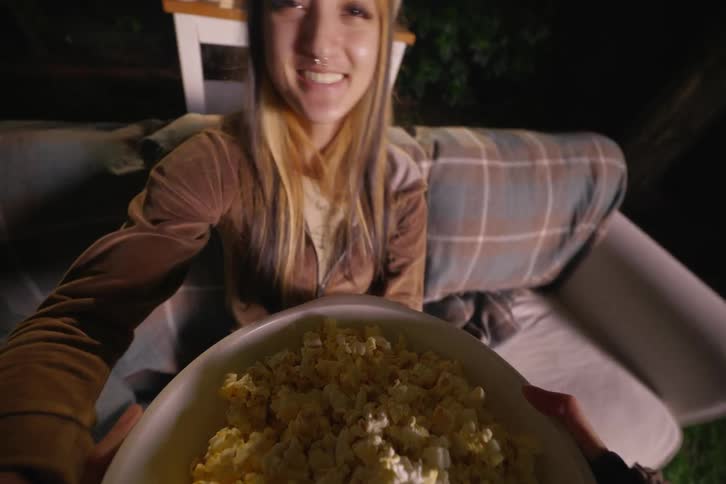 A POV shot of bringing a bowl of fresh popcorn to a smiling friend sitting on a couch for a cozy backyard movie night