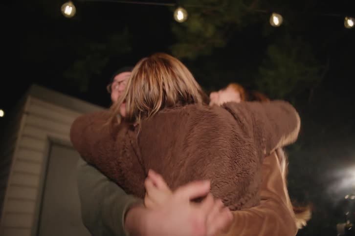 A trio of excited friends leap up from a couch and celebrate wildly while watching a movie in a backyard at night