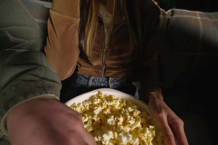 Close up shot of two people's hands reaching into a large bowl of popcorn while sitting on a couch together