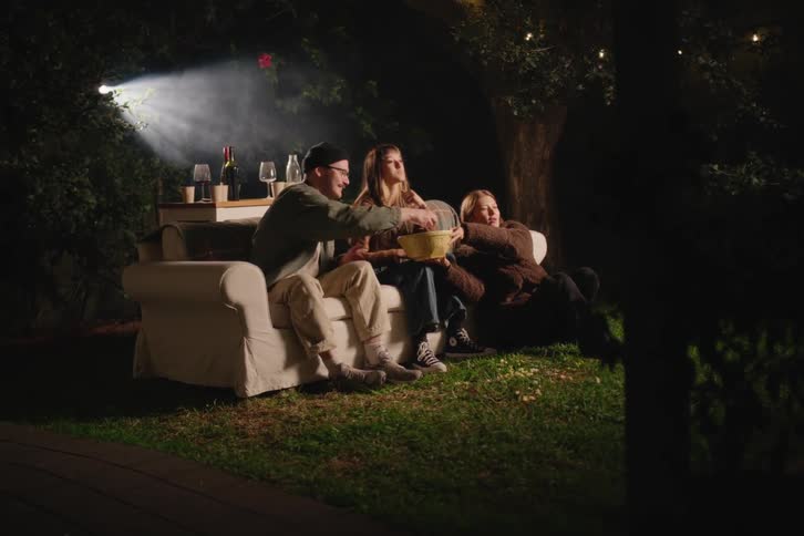 Three happy young friends watch a movie on a projector in their backyard at night, sitting on a couch and eating popcorn together