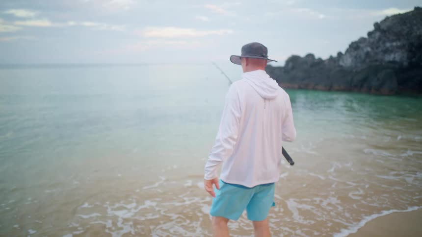 A man in a fishing hoodie and hat walks in slow motion towards the calm ocean from a sandy beach to begin fishing