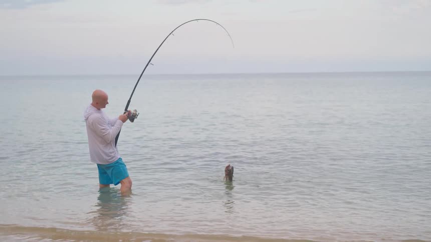 An angler standing knee-deep in the ocean fights to reel in a large fish that is thrashing on the surface of the water