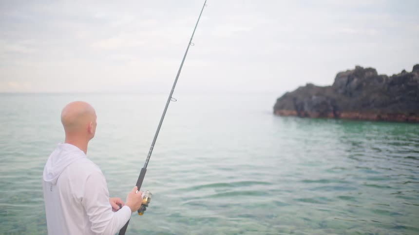 A man patiently stands in the shallow, clear ocean water while fishing on a calm and overcast day