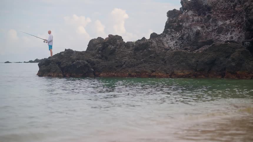 Gentle waves lap the sandy shore as a lone fisherman stands on the rocks in the distance