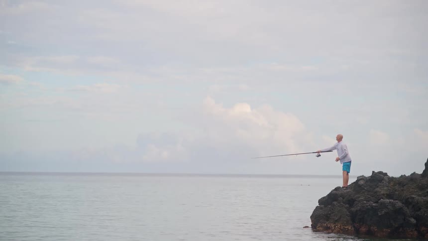 A man casts his fishing rod into the calm sea while standing on rocks along the coast on an overcast day