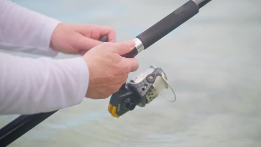 A close-up shot of a fisherman's hands reeling in the line on a fishing rod in clear, shallow water