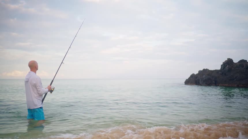 A bald man stands patiently in the shallow ocean water, holding a fishing rod on a calm, overcast day