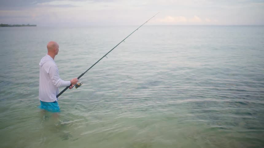 A lone fisherman stands in the calm, shallow ocean waters casting his line during a serene, overcast morning