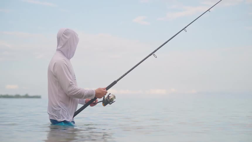 A serene, low-angle shot shows a fisherman in a white hoodie standing patiently in the calm, shallow ocean water