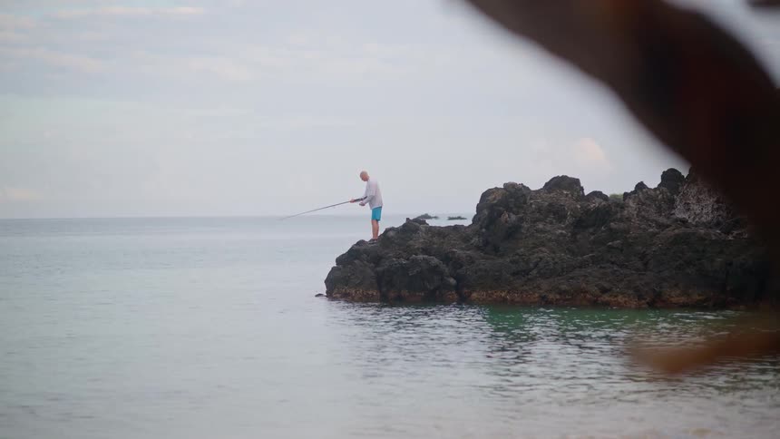 A lone fisherman stands on a rocky outcrop by the shore, patiently casting his line into the calm, overcast ocean