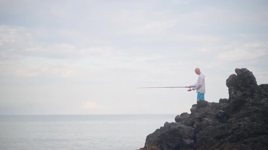 A lone fisherman stands on a rocky outcrop, patiently casting his line into the calm ocean under an overcast sky