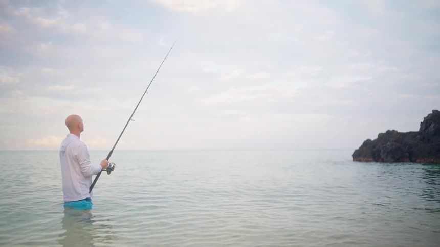 A bald man enjoys a peaceful moment of surf fishing in the tranquil sea on a cloudy day