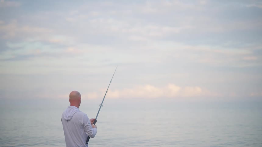 A lone fisherman casts his line into the calm, hazy ocean in slow motion during a beautiful sunrise