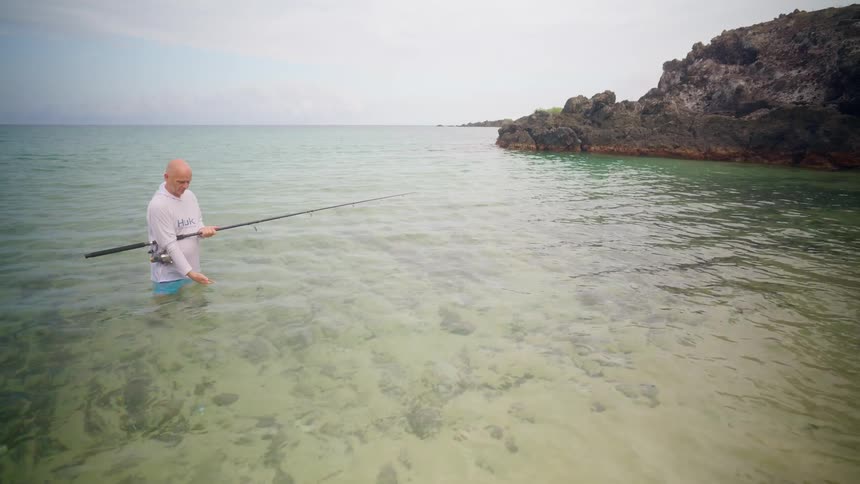 A man patiently fishes in the calm, clear turquoise waters of a tropical ocean cove on an overcast day