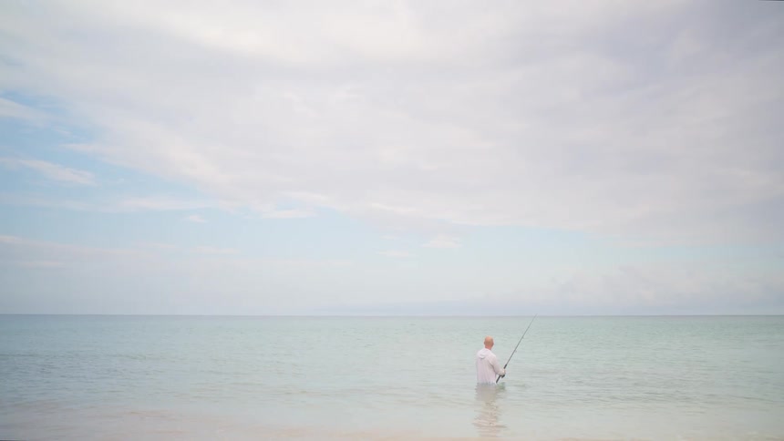 A solitary man patiently fishes in the calm, shallow waters of the ocean under a vast, cloudy sky
