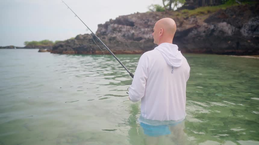 A man wearing a white shirt stands in the clear shallow ocean water while patiently fishing near a rocky shore