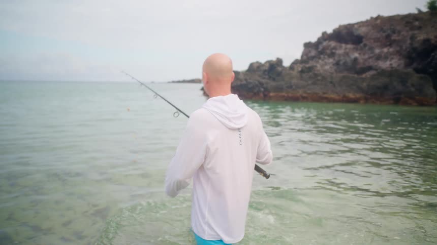 A fisherman wades into the calm, clear ocean from a sandy beach and casts his fishing rod