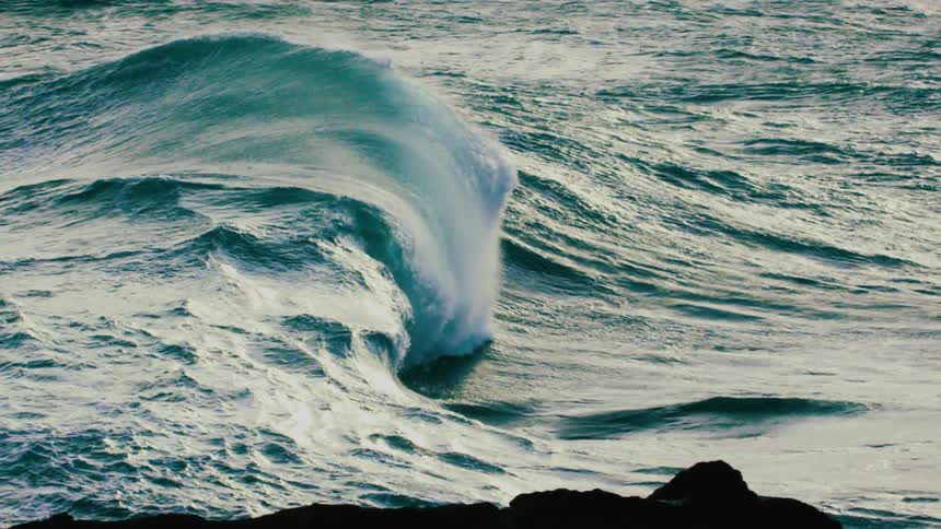 A powerful turquoise ocean wave swells and curls in stunning super slow motion as it approaches a rocky shore