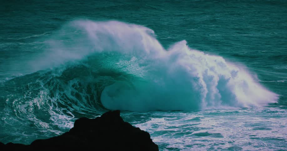 A beautiful and powerful teal wave curls and crashes in mesmerizing slow motion with a rocky silhouette in the foreground