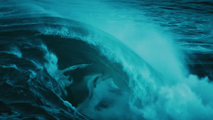 An immense, dark teal ocean wave crests and crashes in dramatic and mesmerizing slow motion