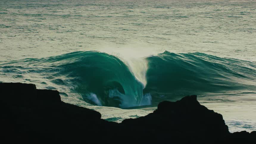 A massive, powerful ocean wave swells and crashes in dramatic slow motion behind a rocky silhouette