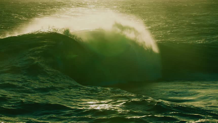 A powerful ocean wave with a green hue crests and breaks in dramatic slow motion as the sun reflects off the water
