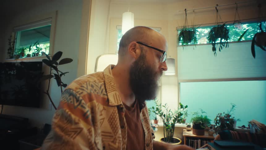 A dedicated bearded man works from home, writing notes and using a laptop in a cozy, plant-filled room