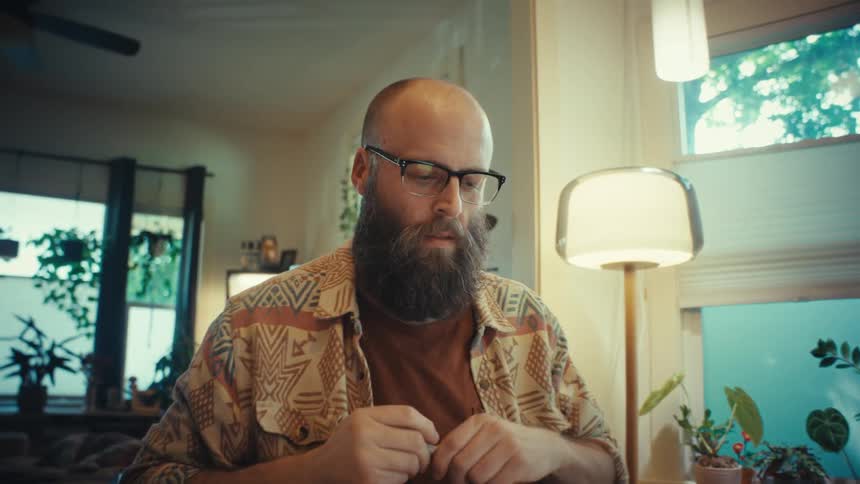 A focused bearded man in glasses studies with a laptop and notebook at a dining table in his plant-filled home