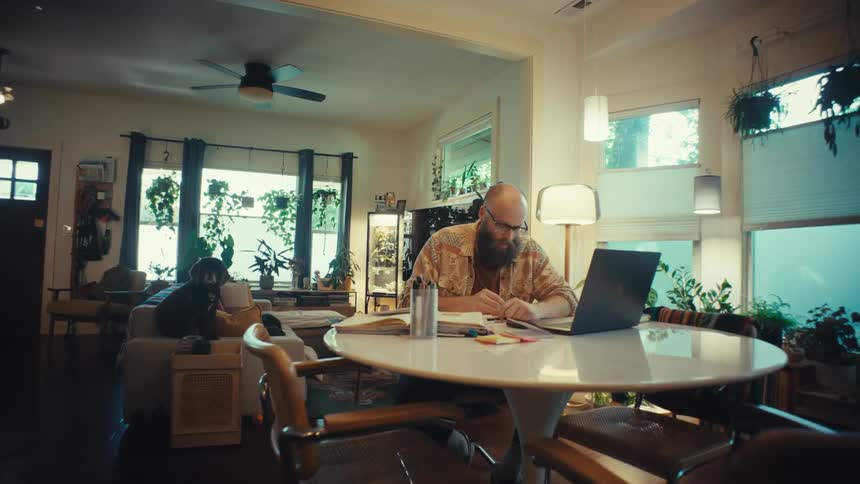 Slow push-in shot of a focused, bearded man working from home on his laptop in a cozy, plant-filled living room while his dog sits on the couch