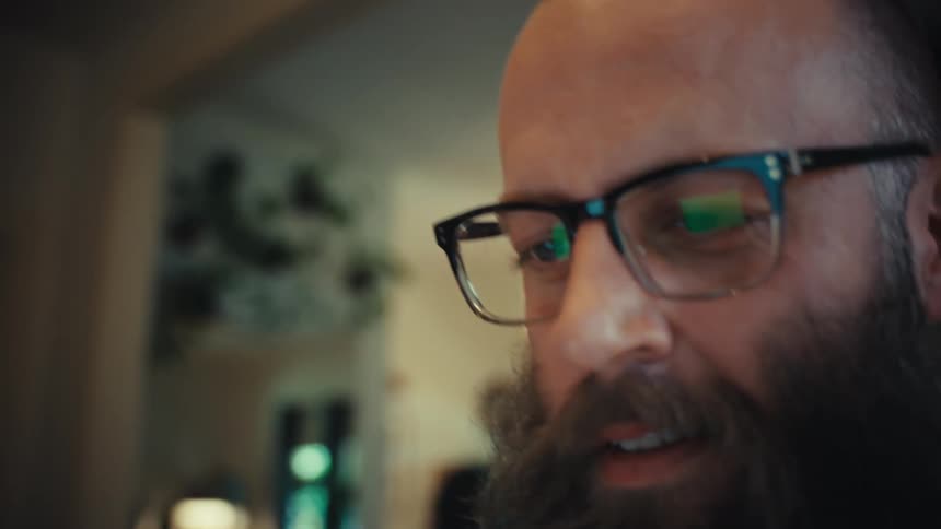 Close up of a man's hands typing on a backlit laptop keyboard, then the camera focuses on his bearded face