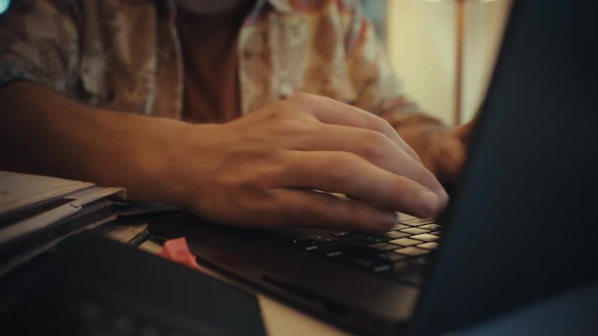 A man with a beard and glasses transitions from writing in a notebook to typing on his laptop while working from his cozy home office