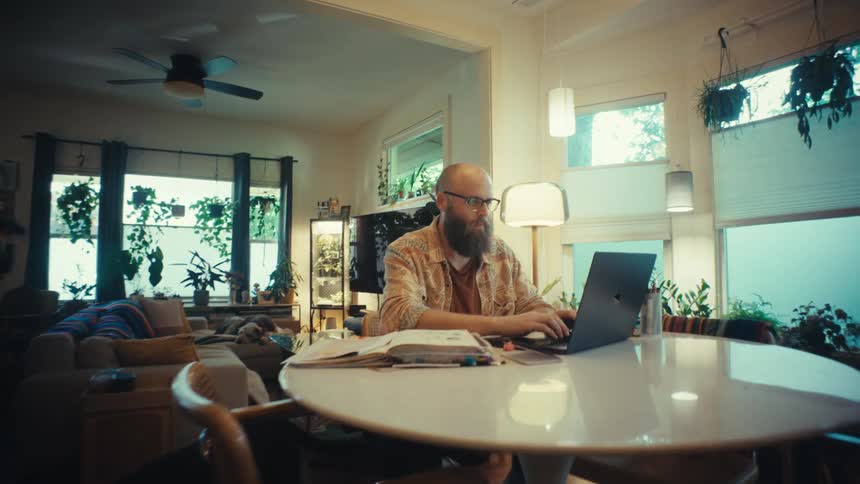 A bearded man with glasses works from home on his laptop in a beautifully decorated apartment filled with plants as the camera slowly pushes in for a close-up