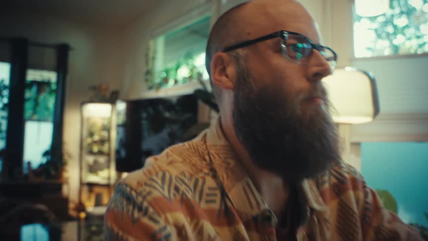 A bearded man with glasses works intently on his laptop from his home office as the camera pushes in for a close-up