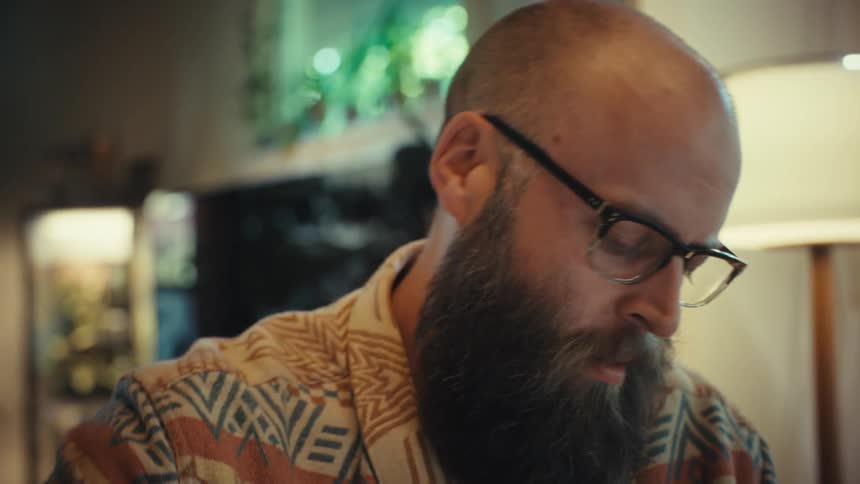 A focused bearded man wearing glasses works intently on a project in his cozy, dimly lit home office