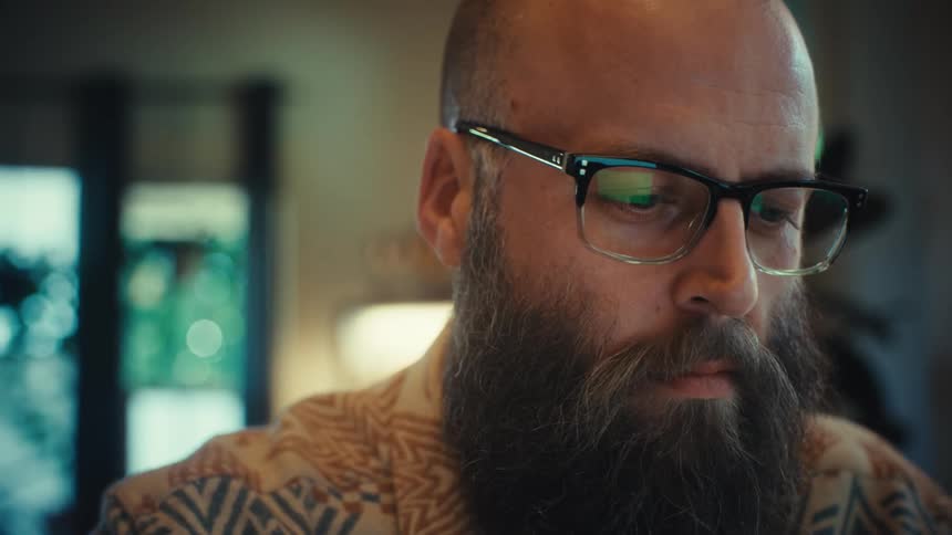 A close-up of a bearded man with glasses focused on typing on his laptop computer