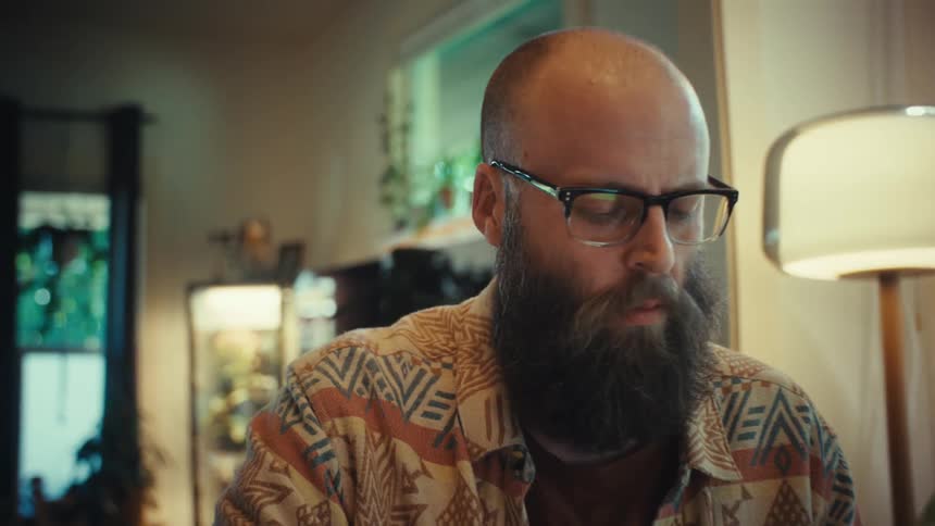 A focused man with a beard and glasses diligently writes notes while studying at his desk in a cozy home office