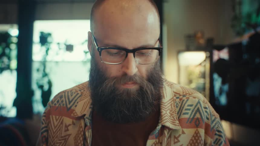 Cinematic dolly-in shot reveals a bearded man with glasses concentrating intently while working from his cozy home office
