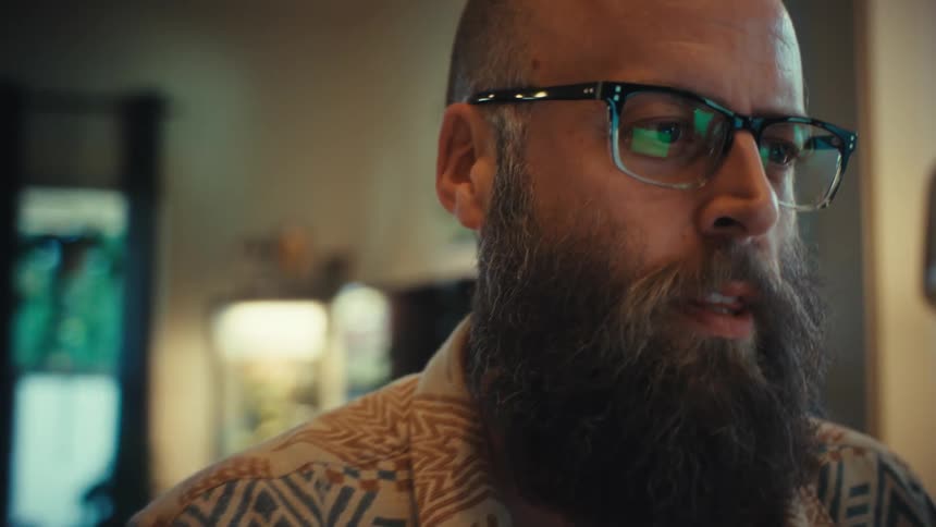 A bearded man wearing glasses is fully engaged in a conversation while looking at a screen in his home