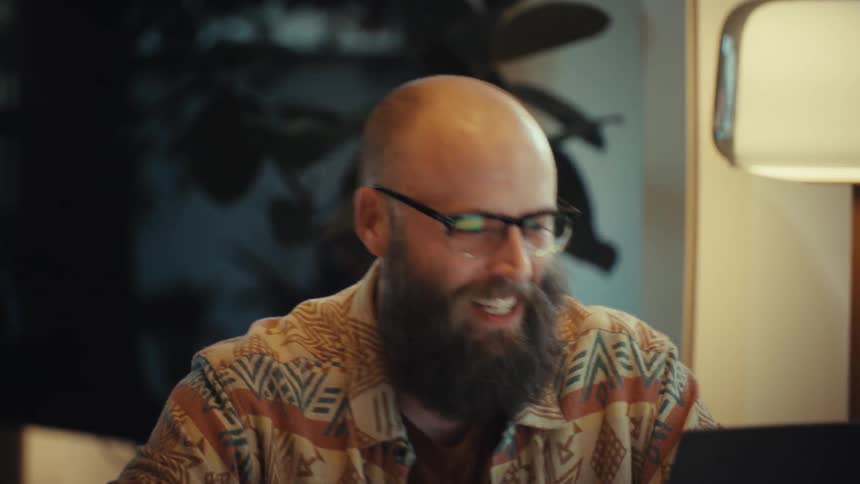 A cheerful bearded man in glasses laughs and claps his hands during a successful video call on his laptop from his cozy home office