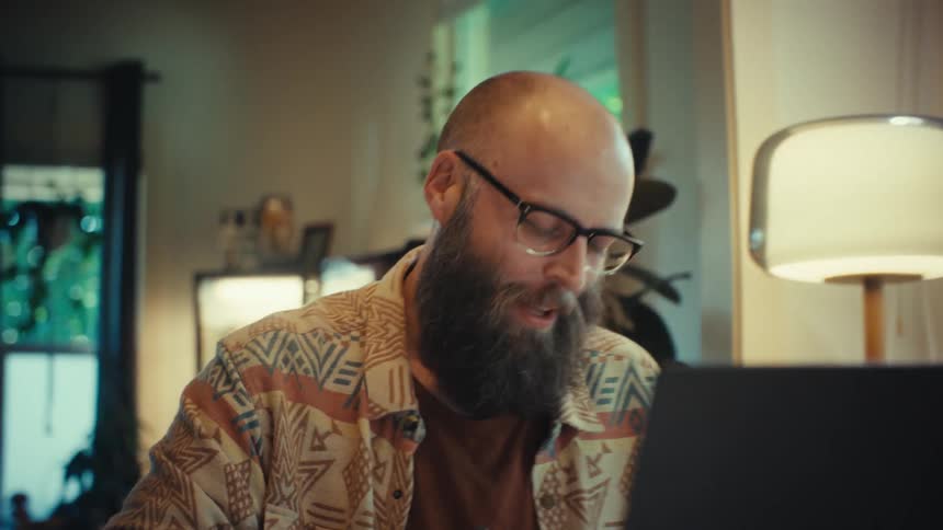 A man with a beard and glasses happily waves during a video call on his laptop while working from his plant-filled home