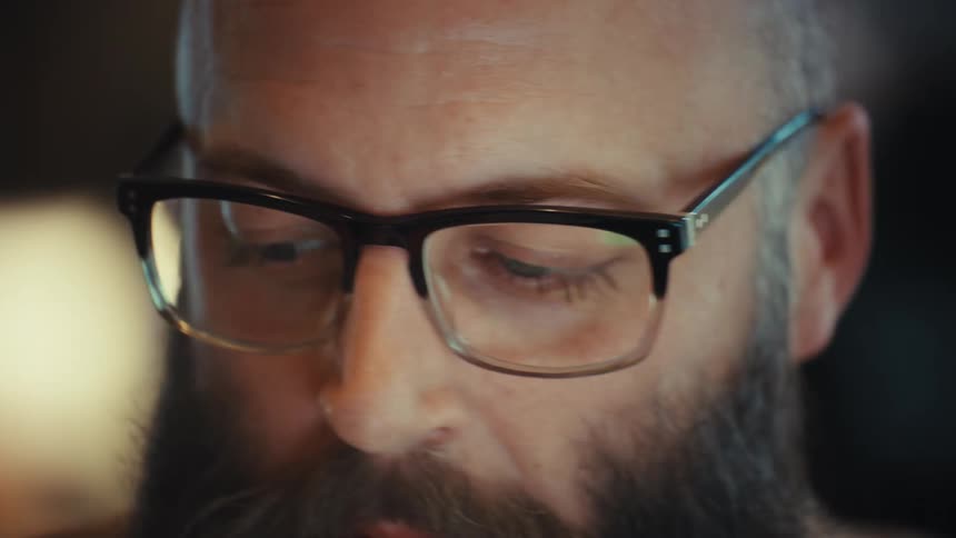 A close-up shot of a bald, bearded man wearing glasses, his eyes intently focused on a screen with light reflecting in the lenses