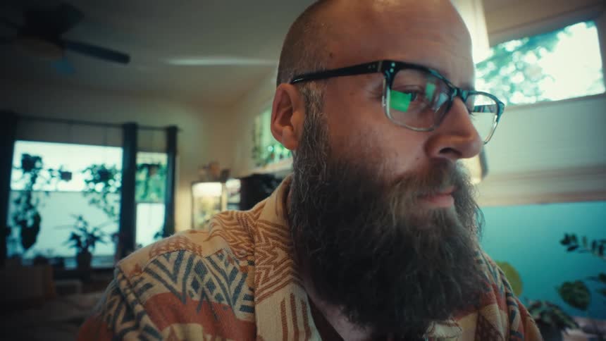 A bearded man wearing glasses types on a laptop keyboard, working intently from his plant-filled home office