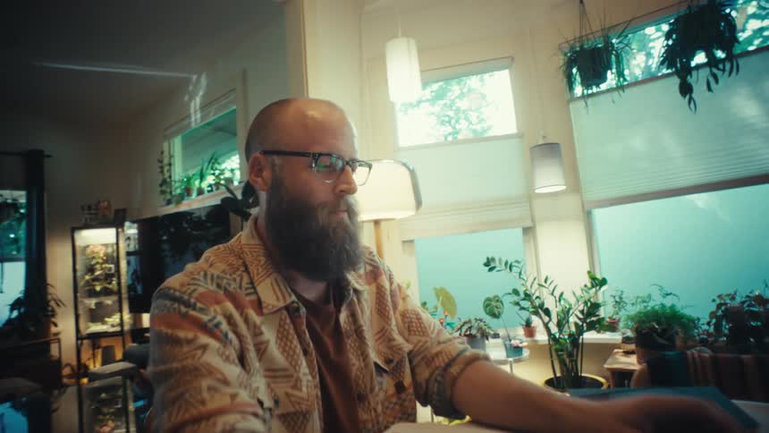 A bearded man opens his laptop to work remotely from his cozy, plant-filled living room