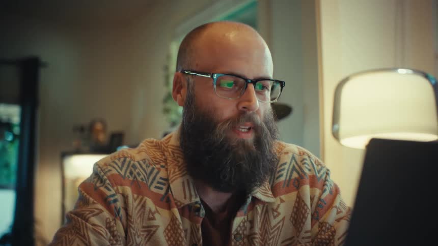 A friendly bearded man with glasses talks enthusiastically on a video conference call while working from his cozy home office